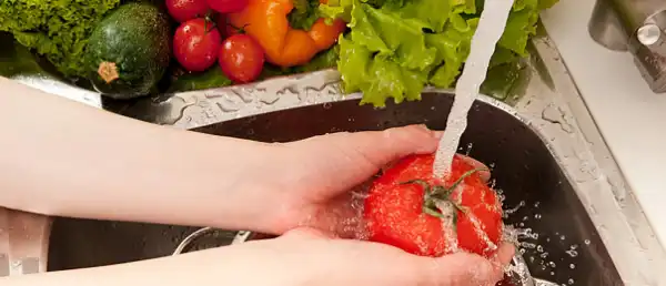 Person's hands rinsing a red pepper in a stainless sink with vegetables sitting on the countertop next to sink.