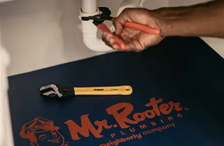 Mr. Rooter plumber's hand fixing under-sink plumbing with a wrench with a branded company mat below the pipes.