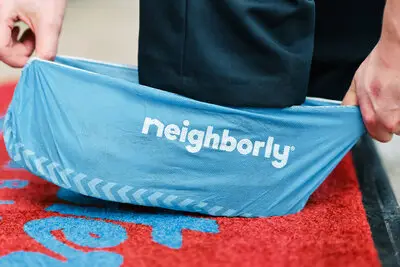 A Mr. Rooter plumber kneeling on a branded doormat and putting on a Neighborly shoe cover