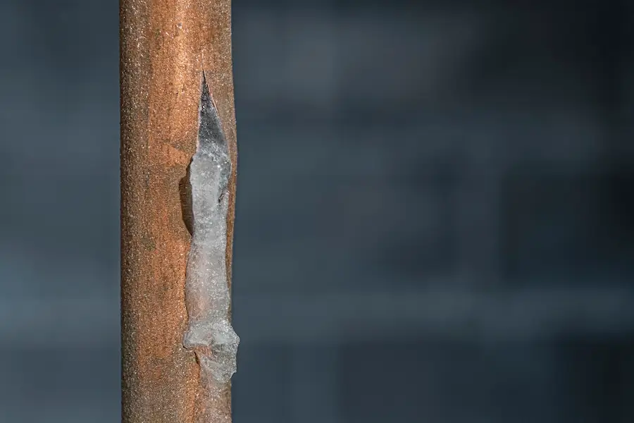 A close-up of a copper pipe that has split from the frozen water inside, that has leaked out and refroze on the outside of the pipe.