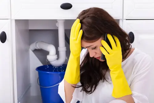 Female customer wearing yellow latex cleaning gloves holding phone with plumbing leak in background.