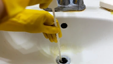A person wearing yellow gloves attempts to clear their sink’s drain using a rubber auger.