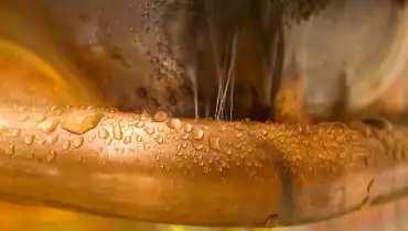 A bent copper pipe covered with droplets from several pinhole leaks that are spraying jets of water.