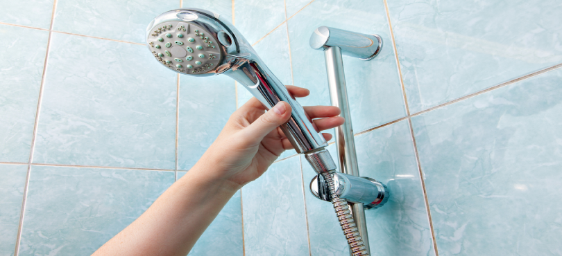 A close-up of a human hand holding a wall-mounted handheld shower head with height adjustable bar slider rail.