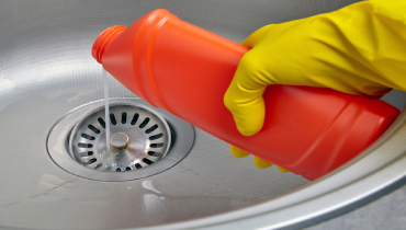 Hand in yellow rubber glove pouring drain cleaner down the kitchen sink drain.
