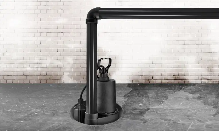 A black sump pump in a basement with concrete flooring and white brick walls.
