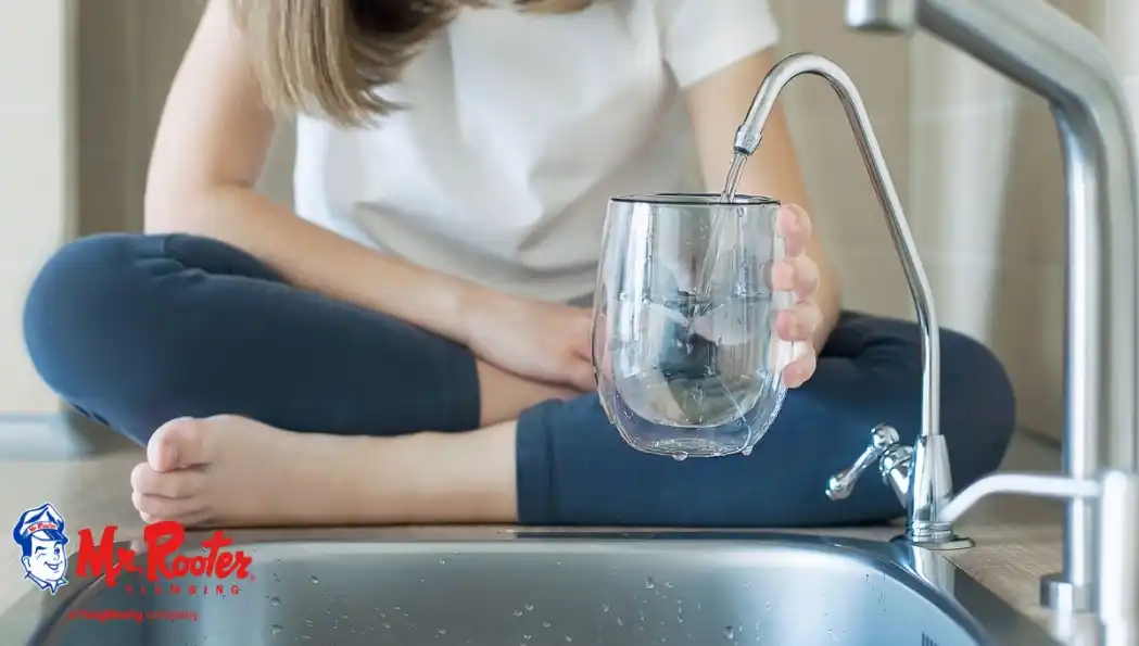 Girl sitting cross-legged on kitchen counter next to the sink, filling up a cup with water from the faucet.