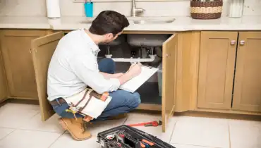 A plumber taking notes while looking under a residential kitchen sink during a plumbing inspection.