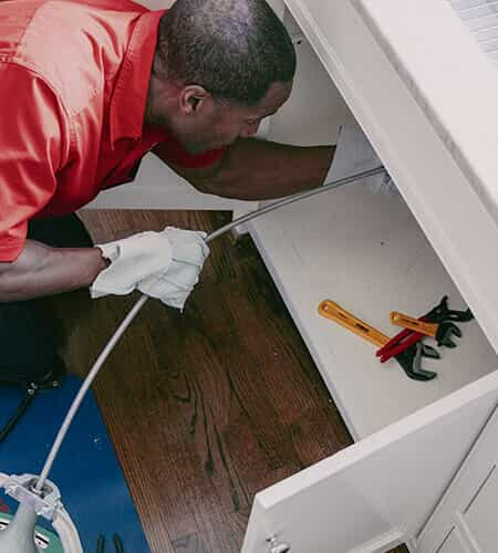 Mr. Rooter plumber crouched next to kitchen sink, using a plumbing snake to clear a clog in a kitchen sink drain.