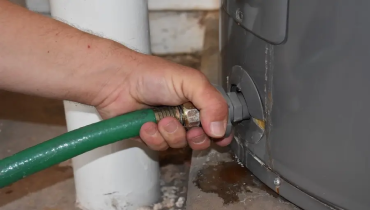 Close-up of a person’s hand attaching a green garden hose to the drain valve at the bottom of a water heater.