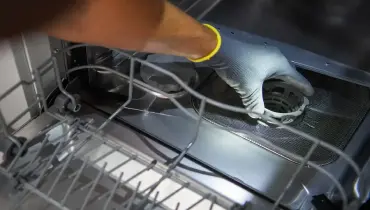 A person wearing a gray glove is removing a filter inside an open dishwasher.