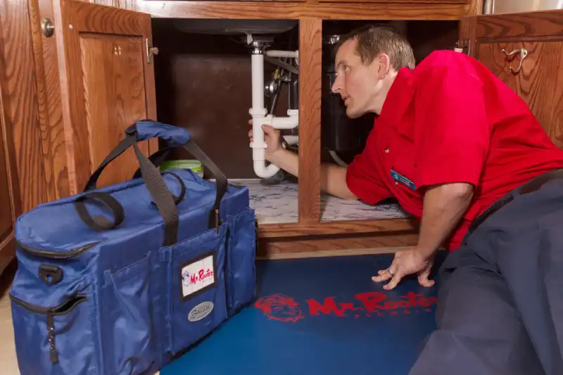 Mr. Rooter plumber leaning on his side to inspect under sink plumbing, with branded company bag and mat next to him.