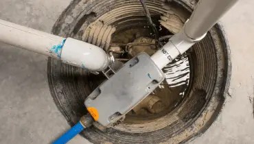 Open sump pit in concrete floor with PVC discharge pipes and fittings over shallow water.