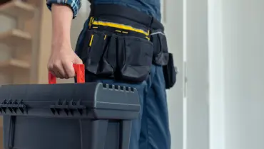 Technician in workwear carrying a black toolbox with a red handle, tool belt visible.