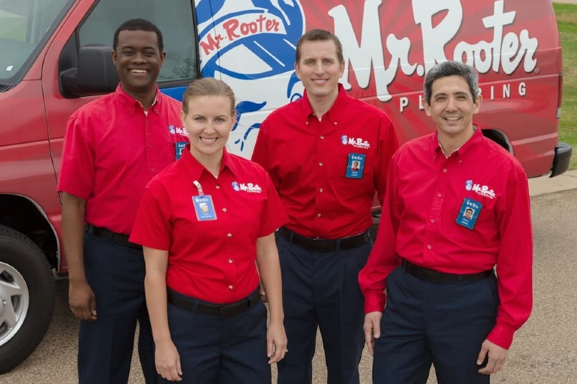 A group of smiling plumbers wearing Mr. Rooter Plumbing uniforms and standing in front of a branded work van.