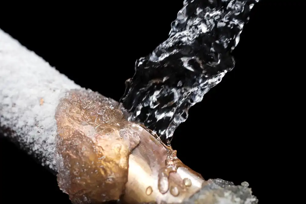 A copper pipe with ice and frost on it, with water streaming out of it of a crack in the side, against a black background.