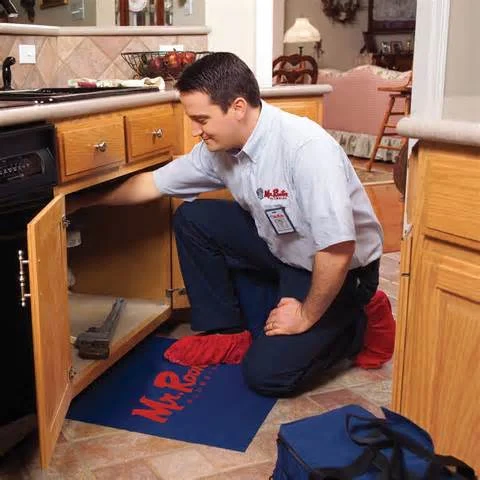 A plumber kneeling on a kitchen floor to inspect and repair a pipe in the cabinet underneath a sink.