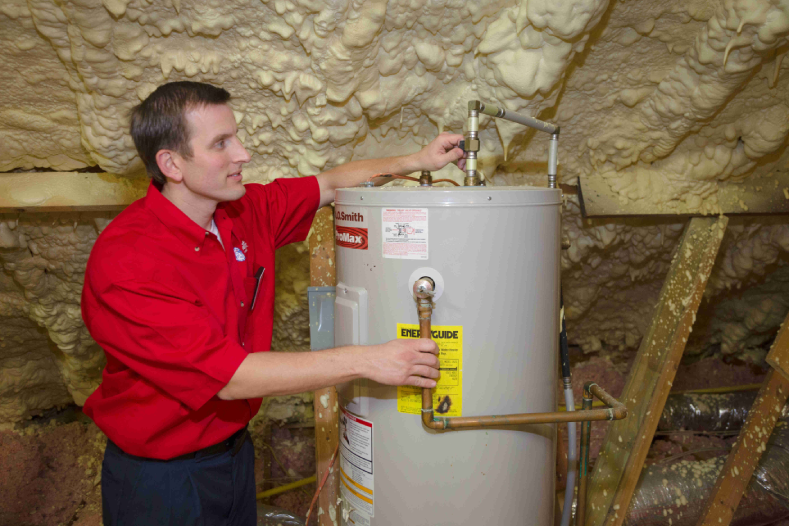 Mr. Rooter service professional repairing water heater in an unfinished interior space with spray-foam insulation visible.