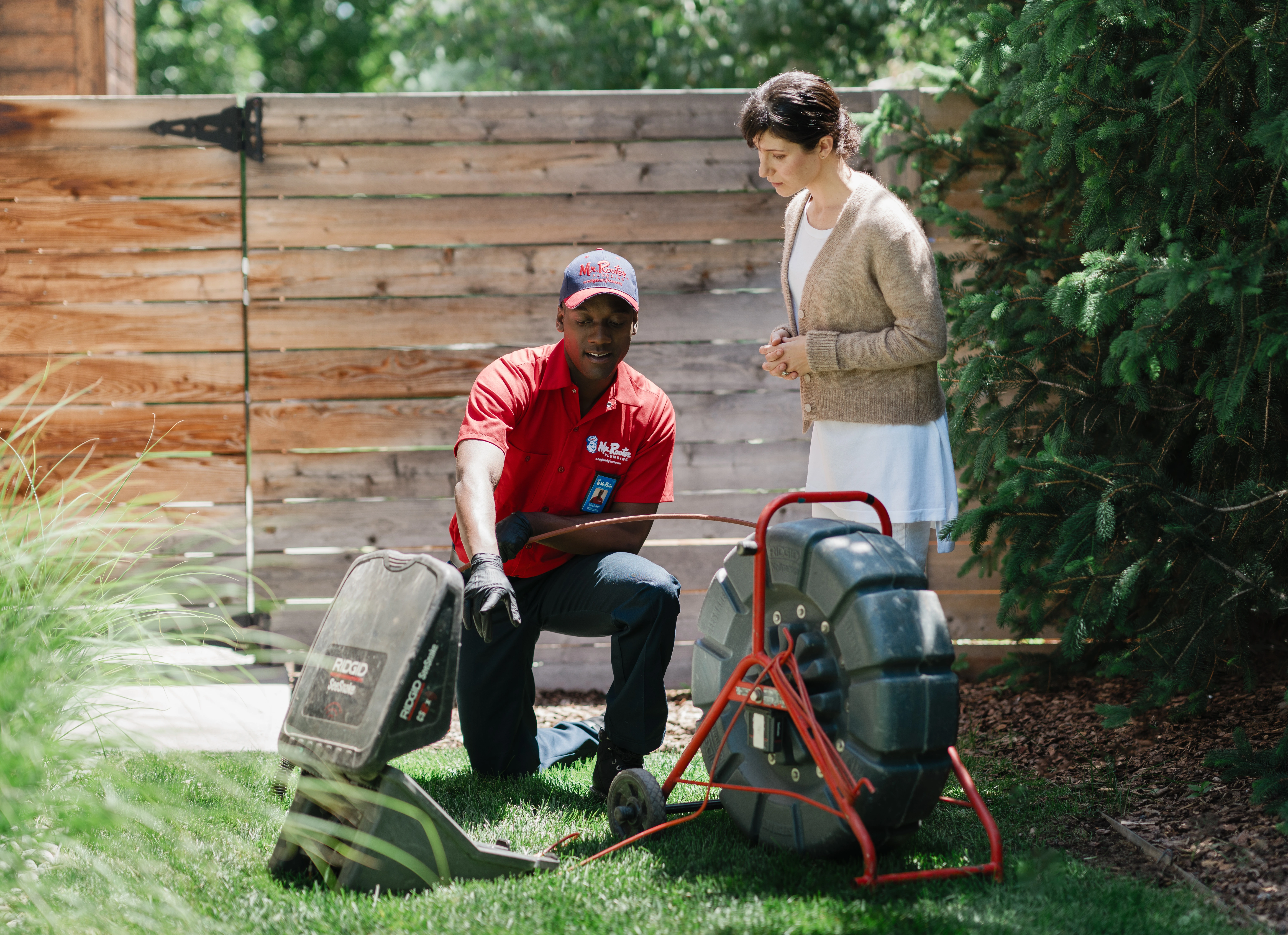 Mr. Rooter Plumbing technician explaining sewer line repair equipment to a homeowner in the yard.