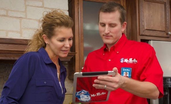 A plumber in a Mr. Rooter Plumbing uniform using an electronic tablet to discuss an estimate with a homeowner.